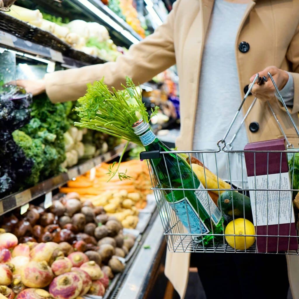 A person shops in a grocery store, holding a basket filled with vegetables, a lemon, and a bottled drink from the produce section.