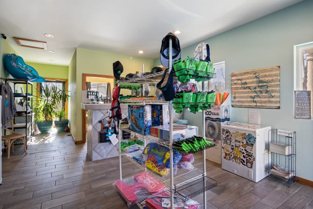 A small, colorful shop interior with various merchandise on shelves, surf gear, plants near the window, and a decorative map on the wall.