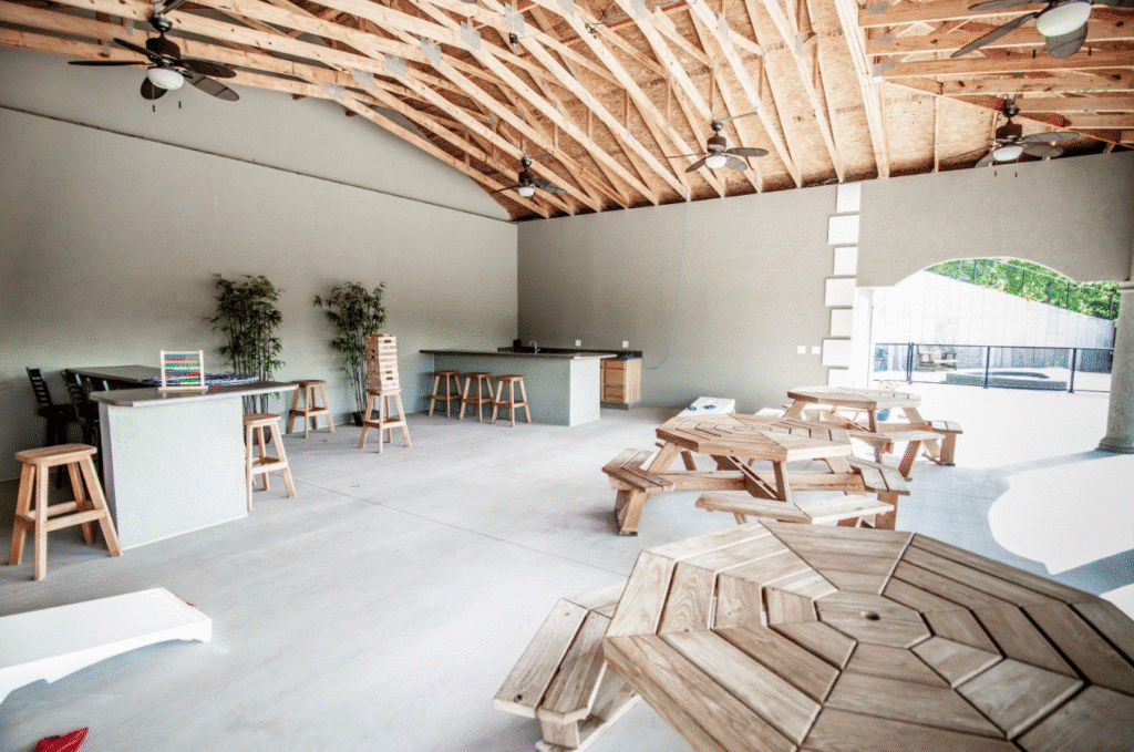 Open-air space with wooden tables, stools, and bar area under exposed-beam ceiling. Outdoor view through archway. Game boards on tables.