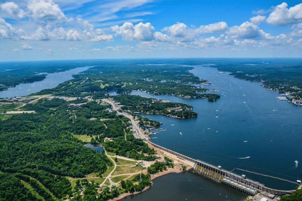 Aerial view of Lake of the Ozarks, displaying lush green forests, winding roads, waterfront properties, boats, and a prominent dam under a partly cloudy sky.
