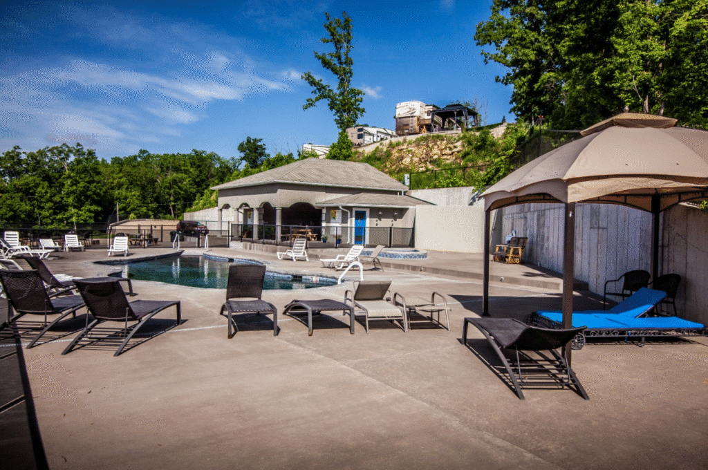 Concrete poolside area with empty loungers, a small building, and a canopy. Trees and parked vehicles in the background under a clear sky.