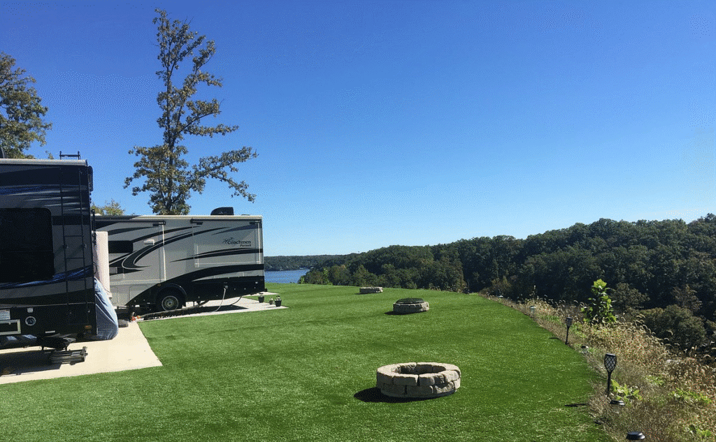 RVs parked on a grassy hill overlooking a lake under clear blue skies. Fire pits are placed near the edge of the hill.