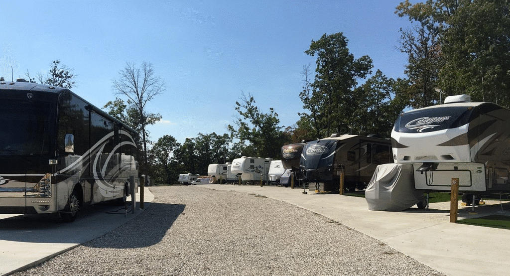 A row of parked RVs and trailers on concrete pads in a wooded campground under a clear blue sky.
