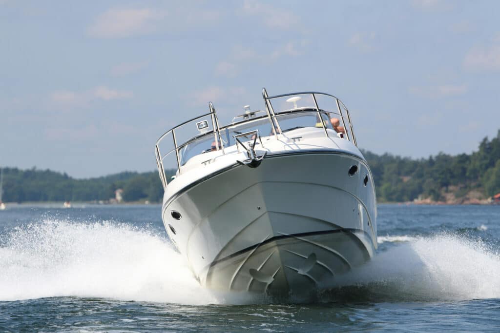 A motorboat with a person on board speeds across the water, creating a splash, surrounded by trees and a blurred shoreline.