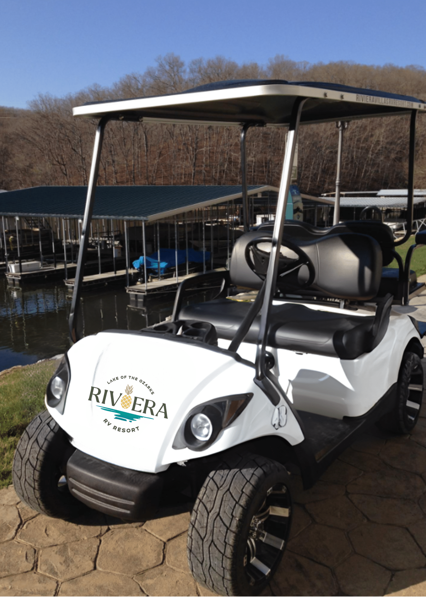 A white golf cart with "Riviera RV Resort" logo is parked near a dock by a wooded lakeside area under a clear blue sky.