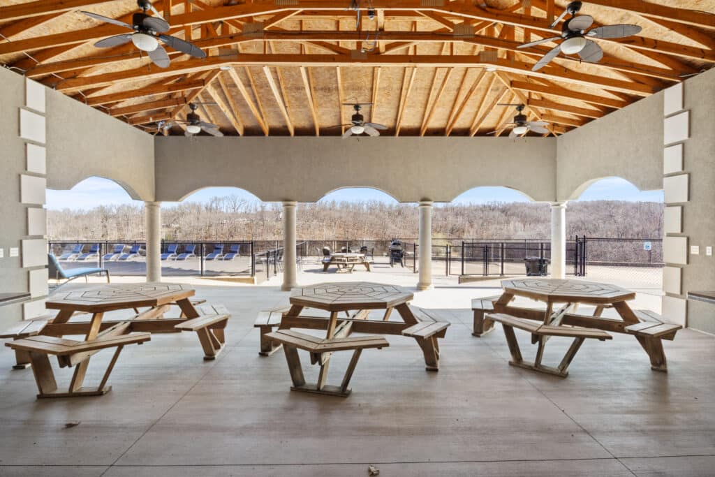Open pavilion with wooden picnic tables, ceiling fans, and arched openings overlooks a fenced outdoor pool area and treeline under a clear sky.