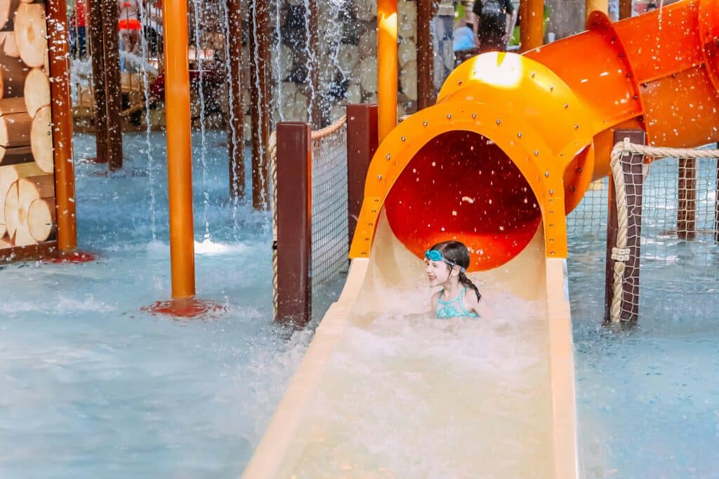 A child wearing goggles slides down a colorful water slide in an indoor water park, surrounded by splashing water and playful structures.