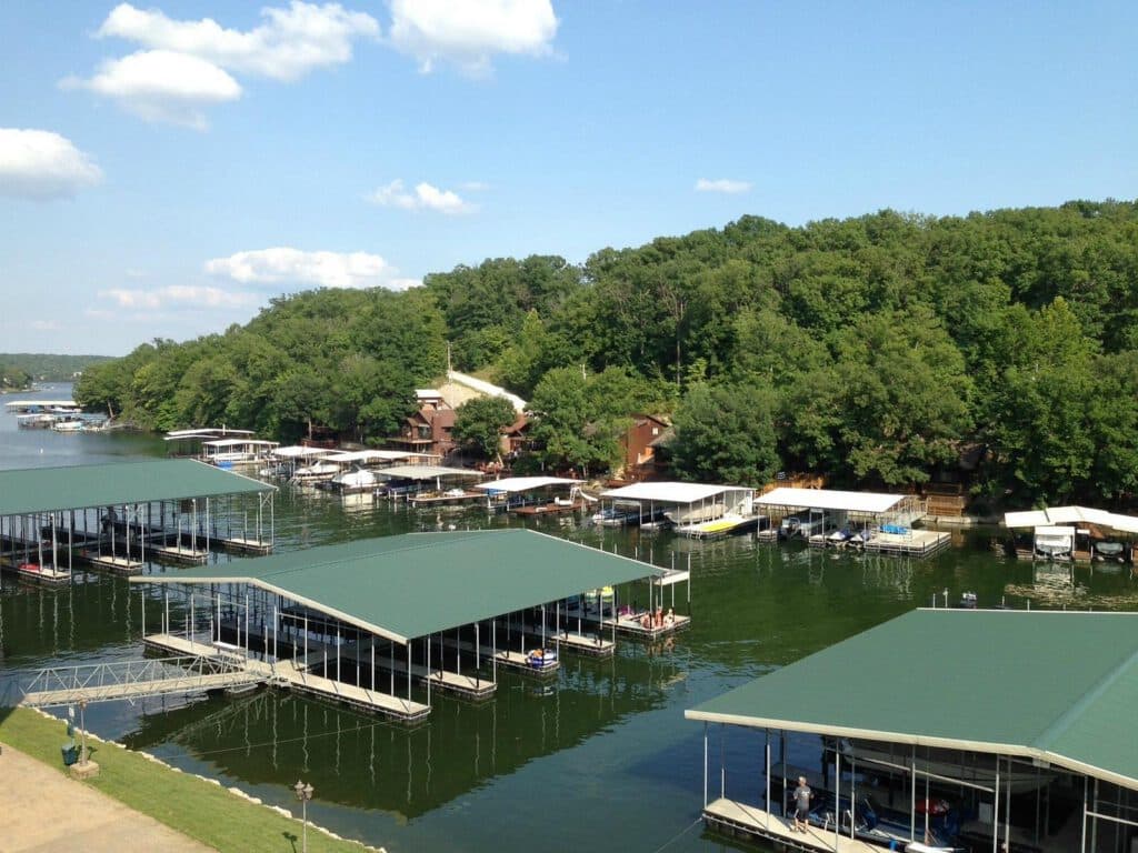 A serene lakeside scene features multiple covered boat docks, surrounded by lush green trees under a blue sky with scattered clouds.