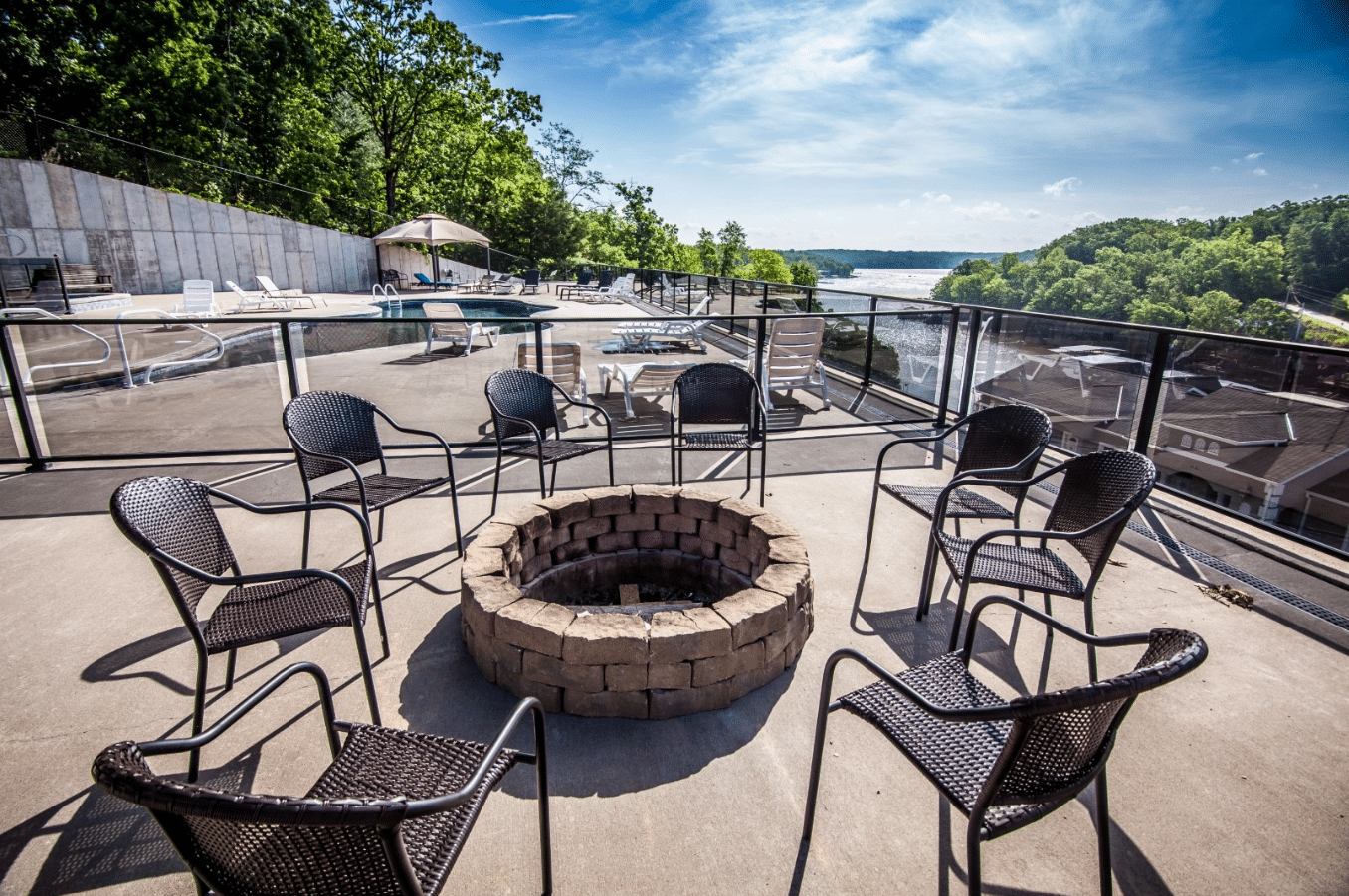 A patio with wicker chairs surrounds a stone fire pit, overlooking scenic views of trees and a river under a clear blue sky.