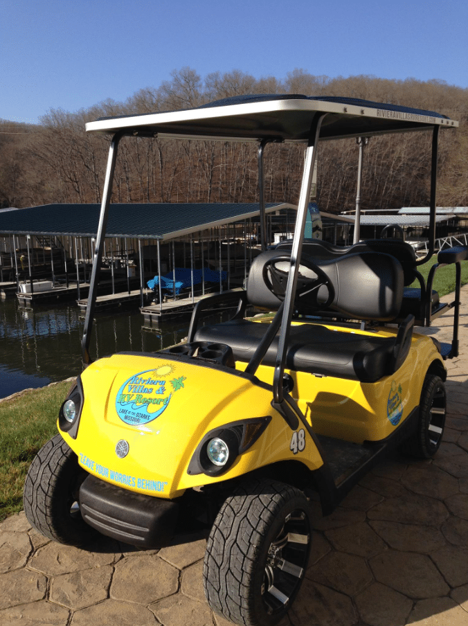 A bright yellow golf cart is parked on a stone path by a lakeside dock, under clear blue sky, amid bare trees.