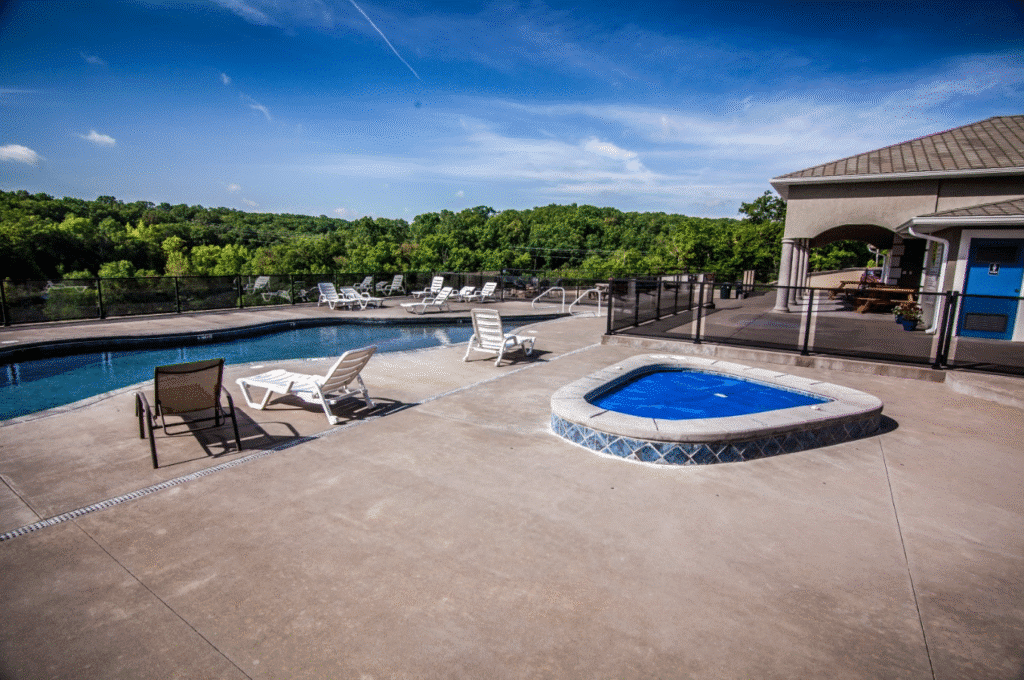 Outdoor pool area with lounge chairs and a nearby building, surrounded by lush green trees under a clear blue sky.