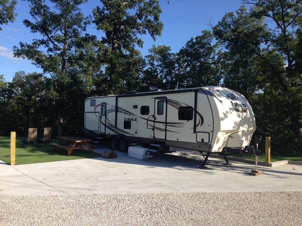 A camping trailer parked on a concrete pad amid trees. Nearby picnic table and fire pit under a clear blue sky.