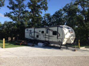 A camping trailer parked on a concrete pad amid trees. Nearby picnic table and fire pit under a clear blue sky.