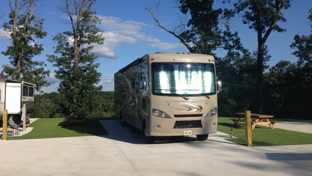 A large RV is parked on a concrete pad near a picnic table, surrounded by trees under a clear, sunny sky.