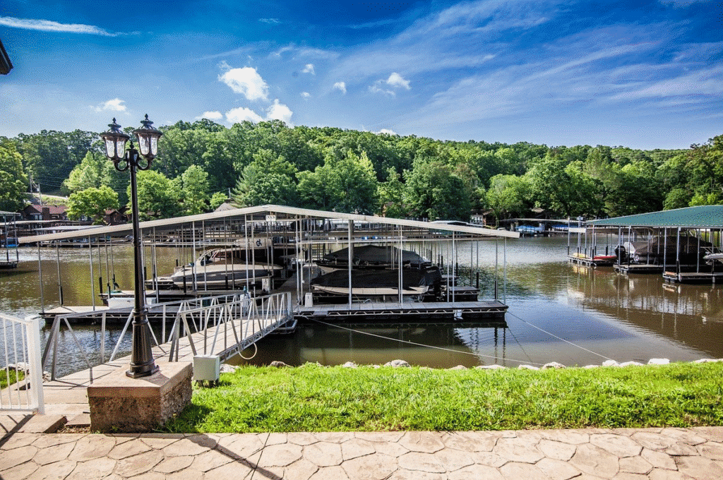 A tranquil lakeside marina with covered boat docks, nestled against lush green trees and clear blue skies, and a decorative lamp post nearby.