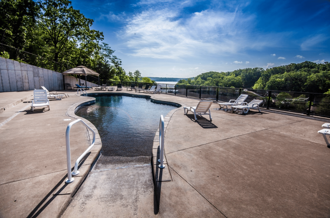 Poolside view with lounge chairs under a clear sky. Trees surround the area, overlooking a river or large lake. Peaceful atmosphere.