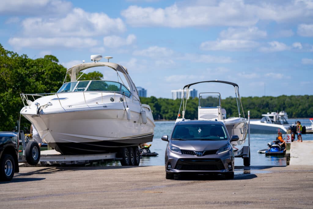 A large boat is towed near a dock with a vehicle. People interact on the water beneath a sunny, blue sky.