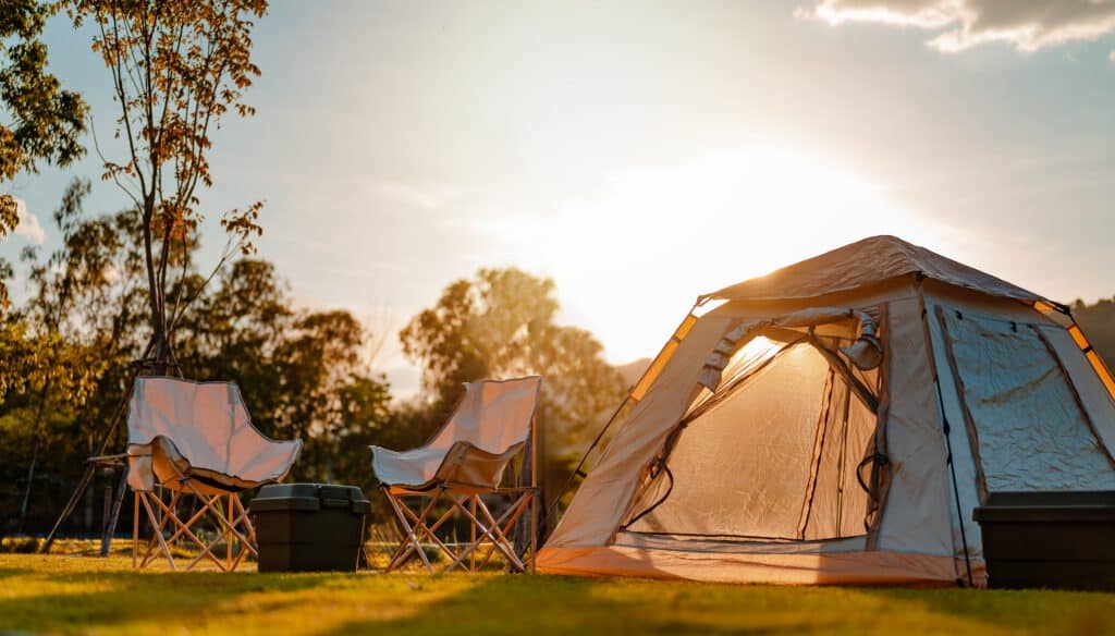 A sunlit campsite with a glowing tent, two empty chairs, a box, and scattered trees in a peaceful outdoor setting.