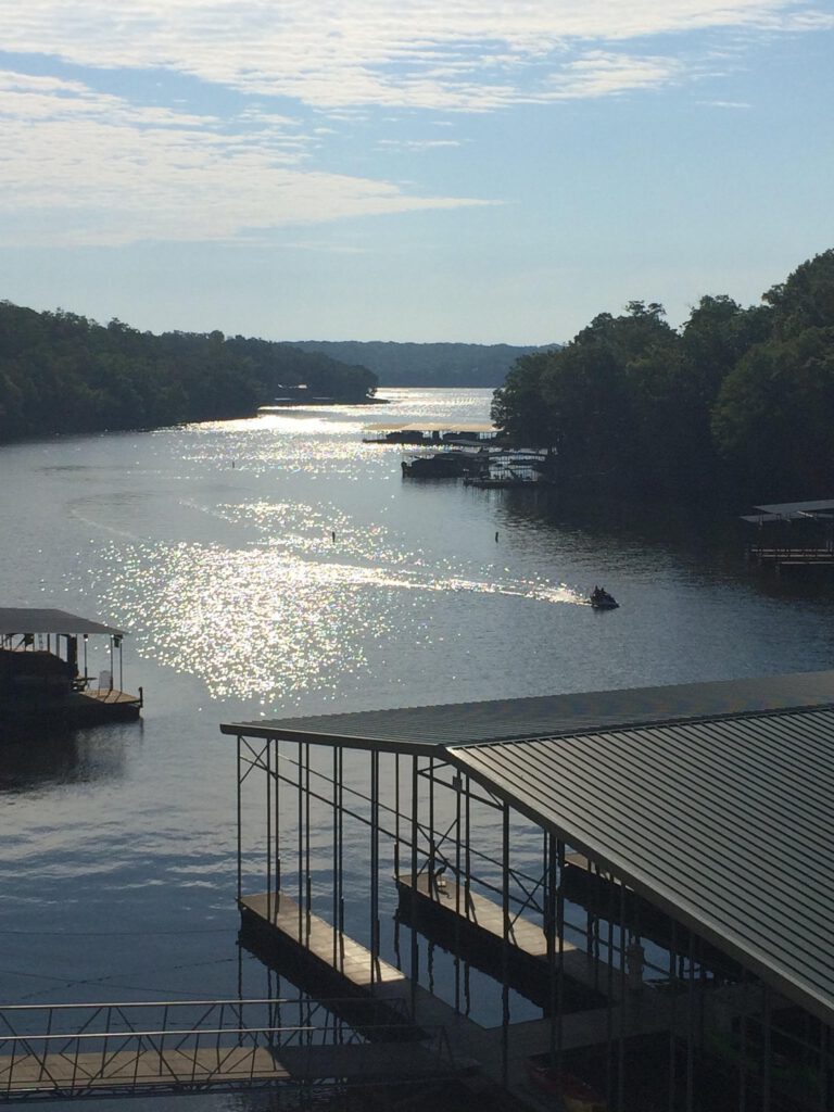 A serene lake view features sparkling water, a dock with covered boat slips, and a person riding a small watercraft under a cloudy sky.